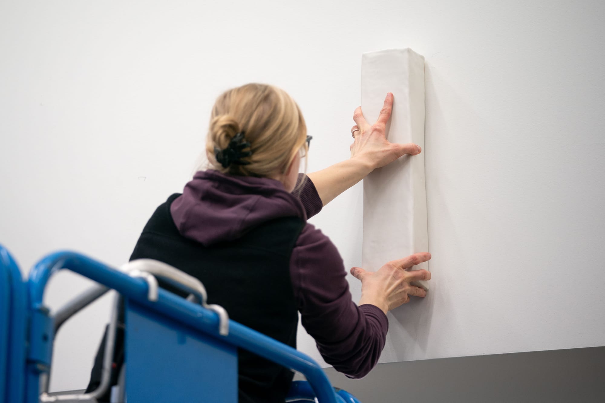 A woman, seen from behind, places a rectangular piece of ceramic pottery on a wall at a high school.