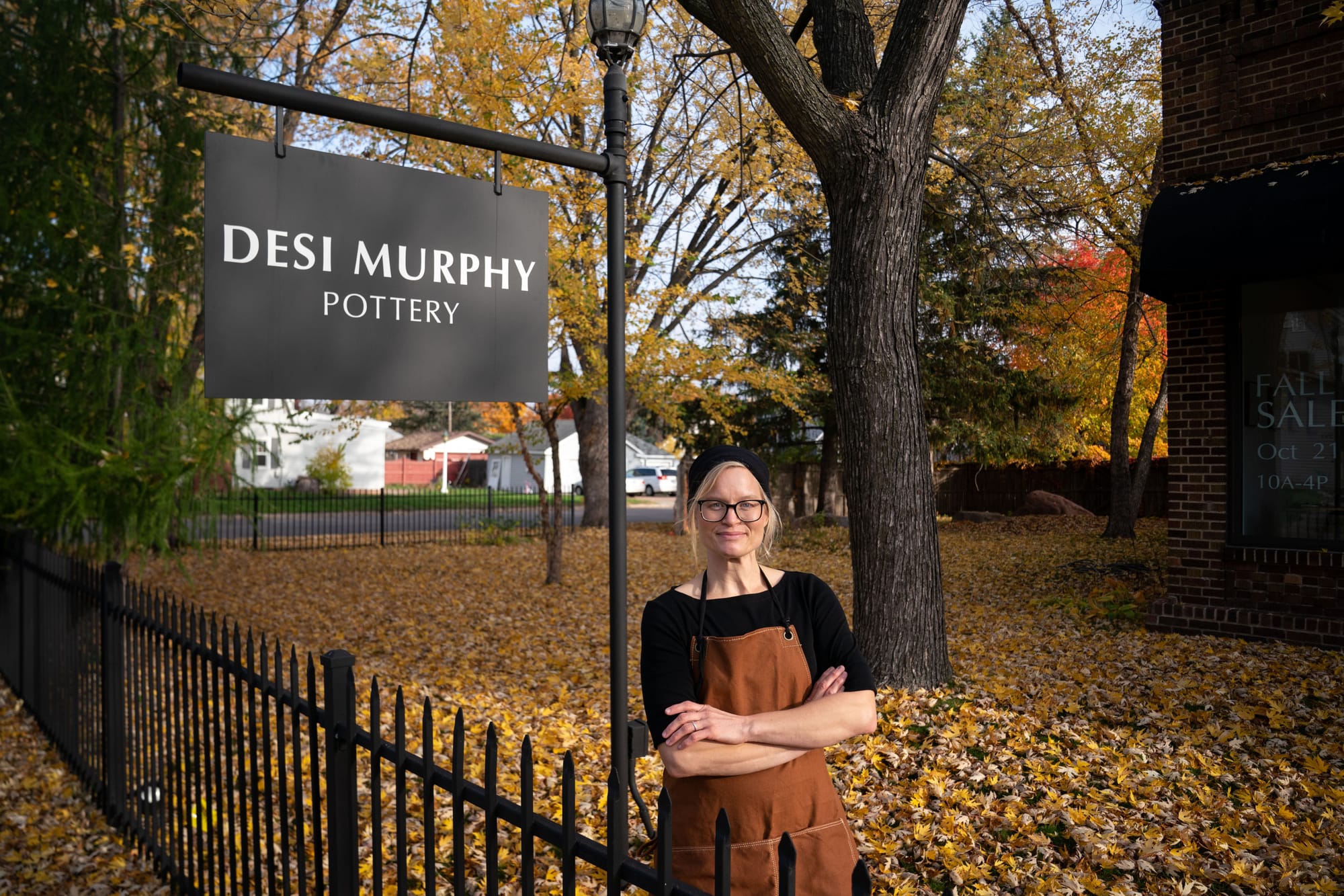 An artist poses for a portrait outside of her studio. The sign is seen in the background.