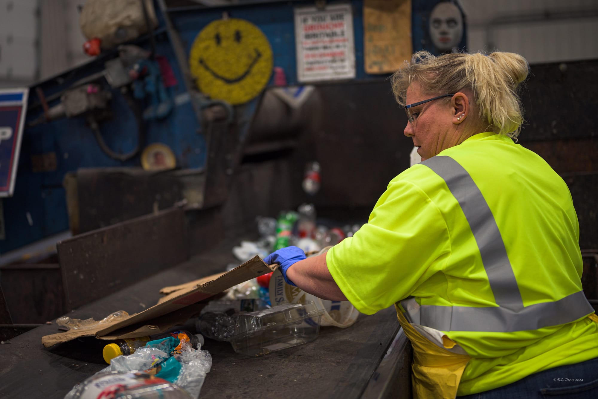 A woman in a high-visibility shirt removes a piece of cardboard from a conveyor belt filled with plastic waste at a recycling sorting facility.