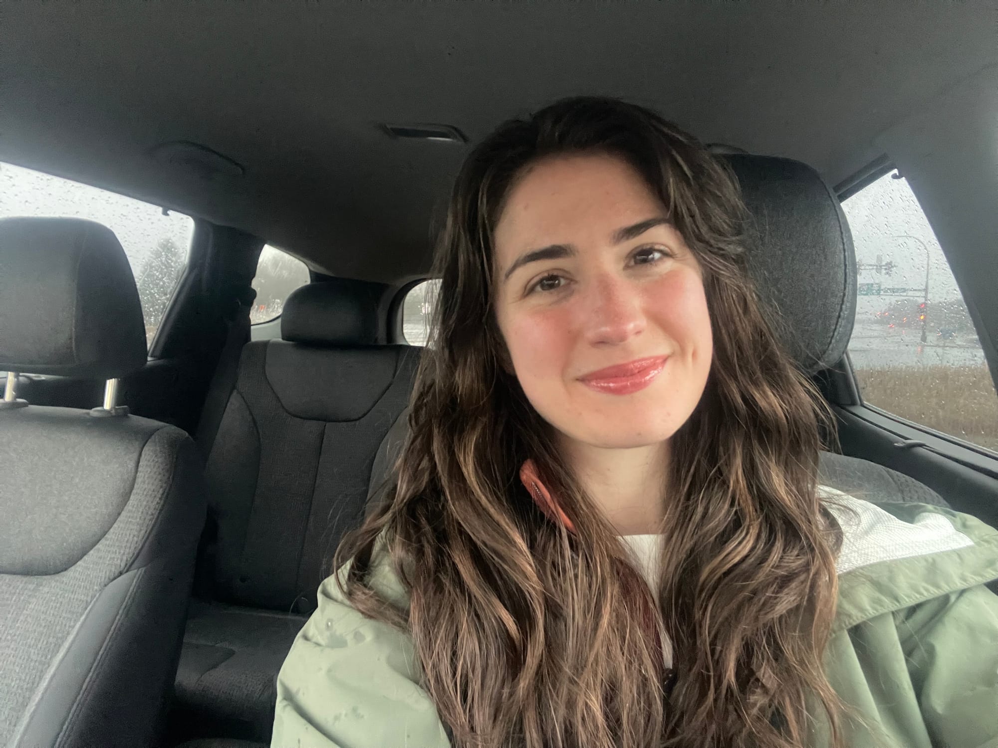 A woman with long, dark, curly hair smiles while she sits in her vehicle. She wears a light green raincoat over a cream colored shirt.