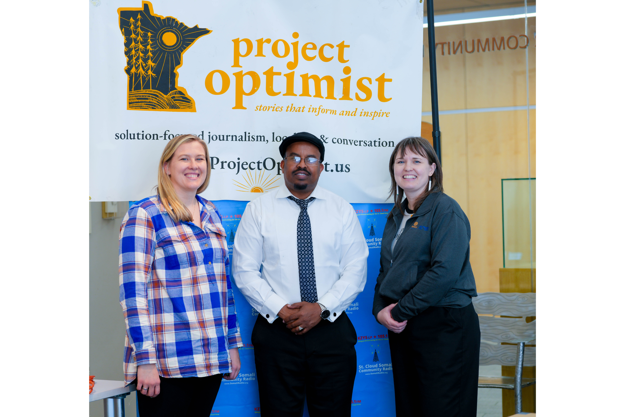 Three people pose for a photo in front of their news organization's banners. There are two women and one man. 