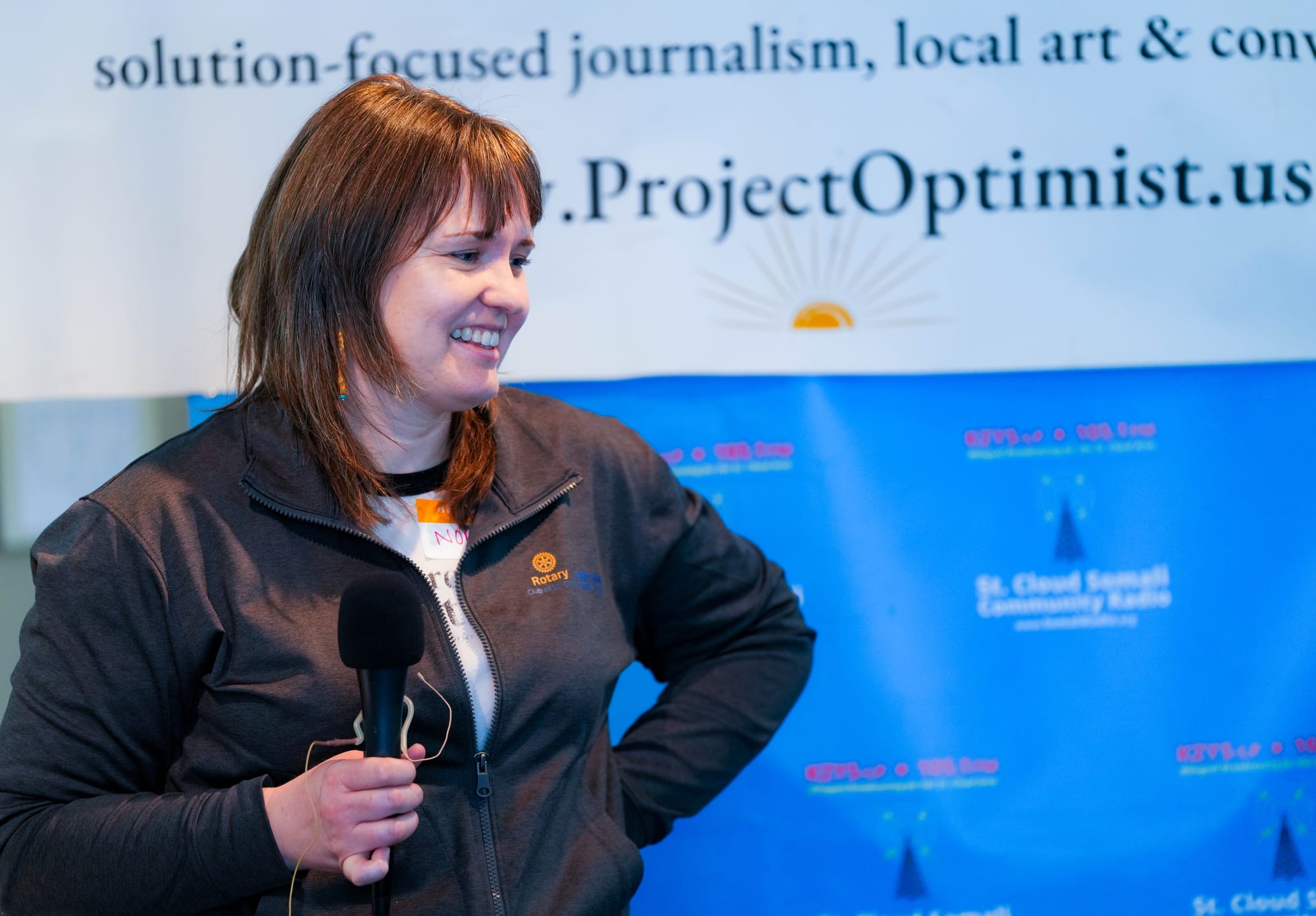 A woman stands in front of banners for Project Optimist and St. Cloud Somali Community Radio. She smiles while holding a microphone during a presentation on journalism. She wears a white t-shirt underneath a black Rotary sweatshirt. 