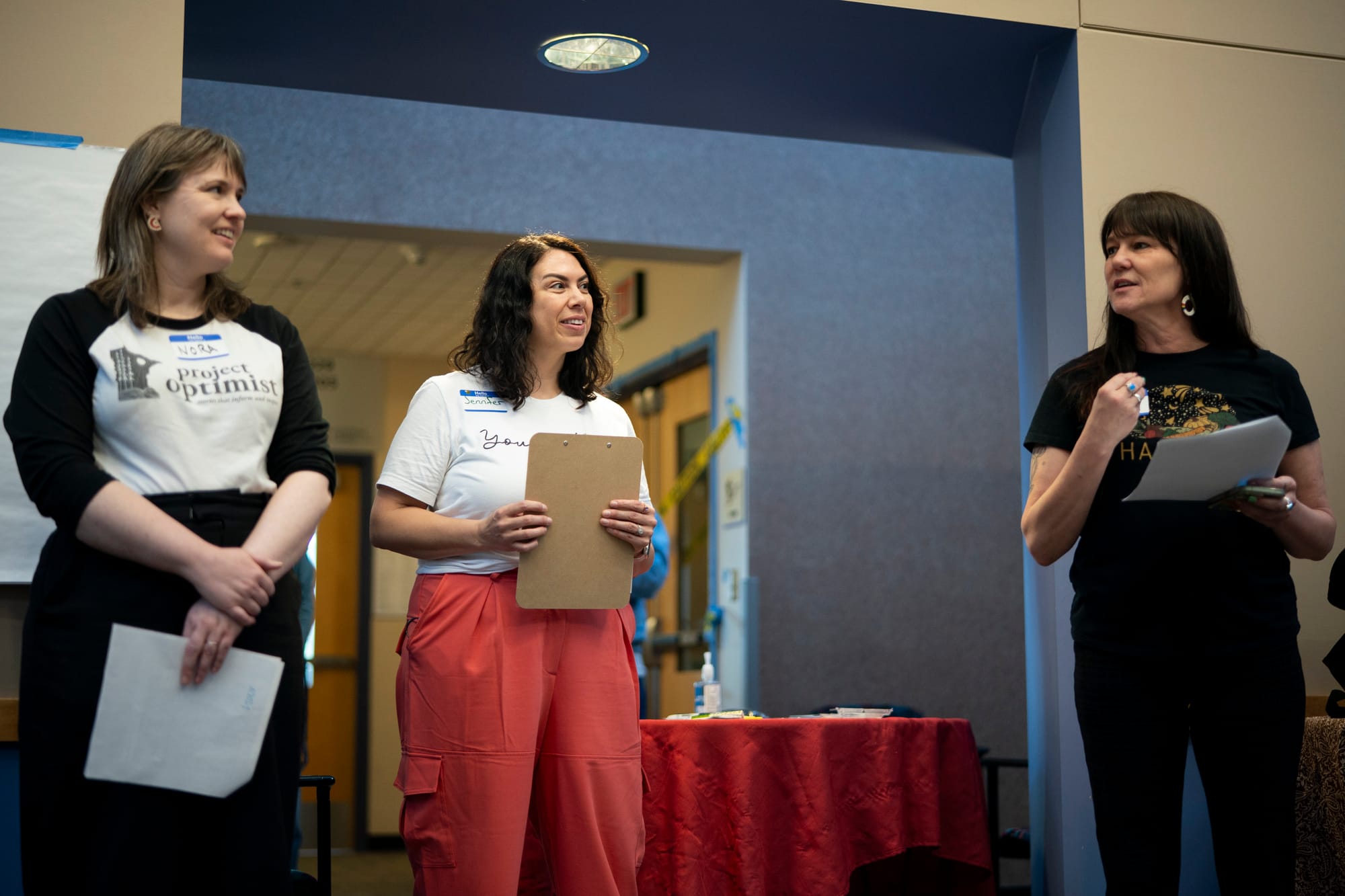 Three women introduce themselves to a group. The woman on the left wears black pants and a black and white T-shirt that says "Project Optimist" on it. She has brown hair. The woman in the middle wears reddish orange pants with a white T-shirt that says "You Matter." She has brown curly hair. The woman on the right wears black pants and a black T-shirt. She has long dark hair. 