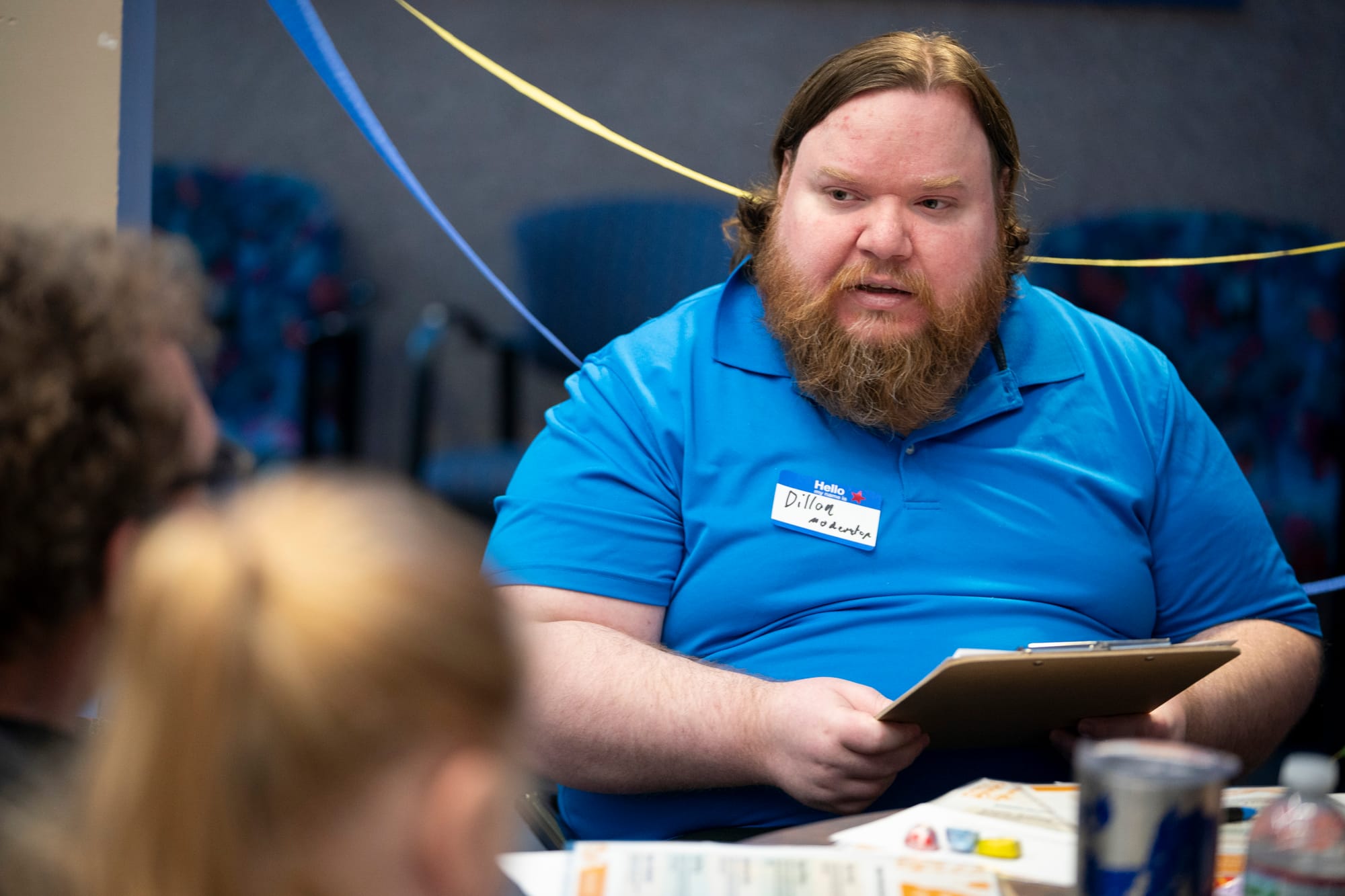 A man asks a question during a moderated discussion event. He wears a bright blue shirt and he holds a clipboard in his hands. He has a red beard and red hair. 