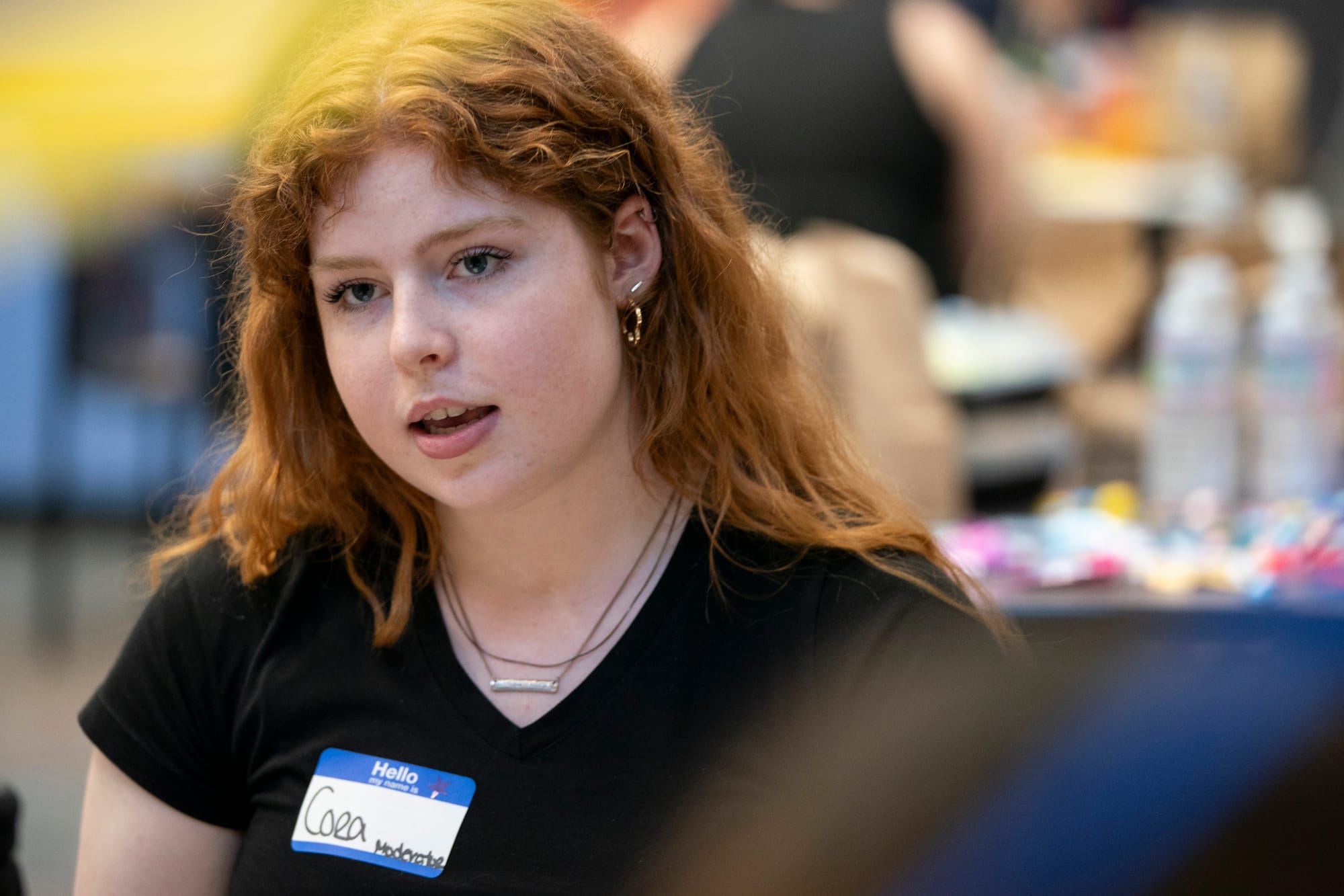 A young woman speaks during a moderated dialogue event. She wears a plain black T-shirt with two black and silver necklaces. She has long, wavy, red hair. 