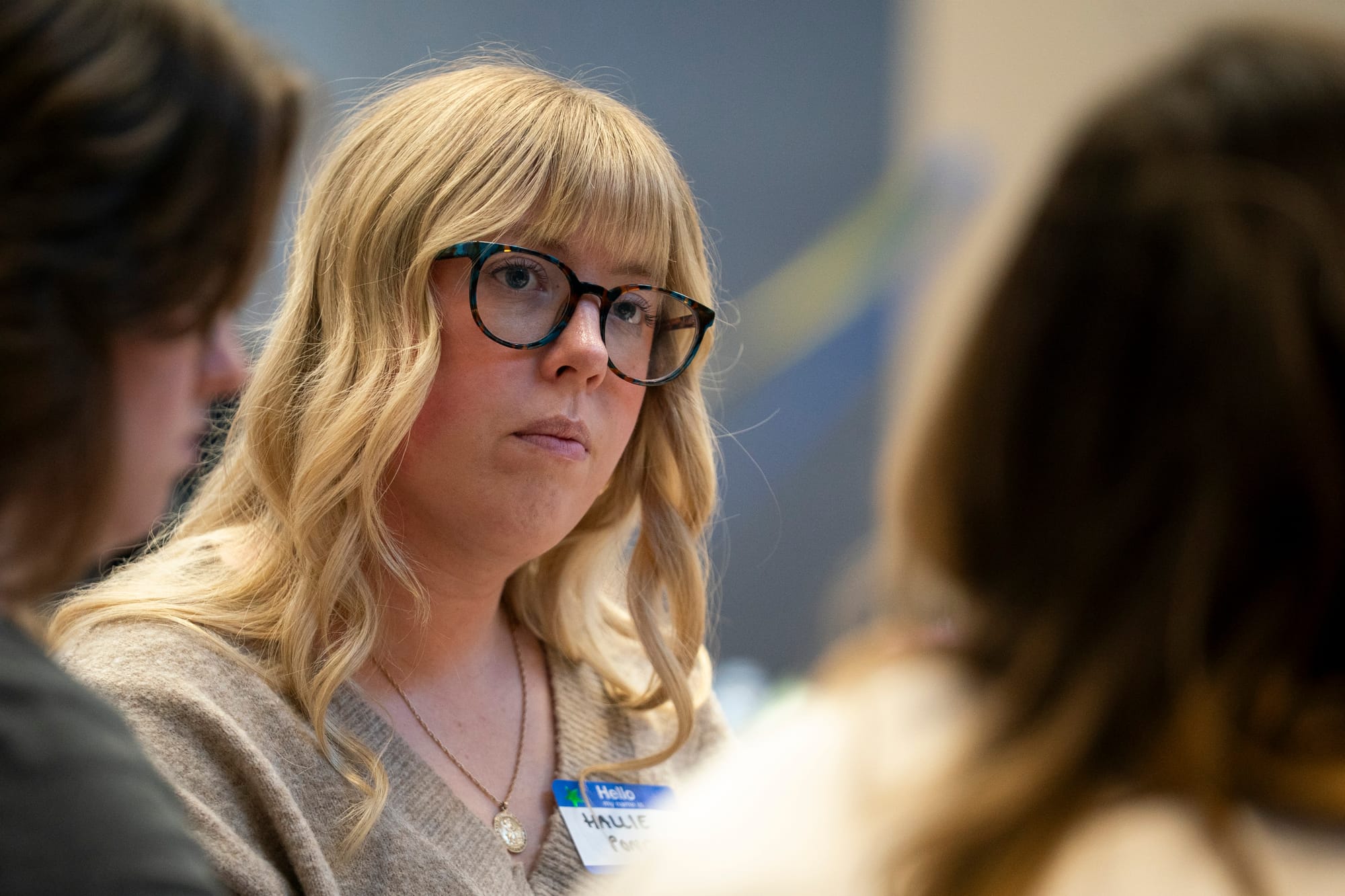 A woman listens carefully while someone else speaks during a moderated discussion. She has wavy blonde hair that falls past her shoulders. She wears a light brown sweater, a gold necklace, and colorful glasses.