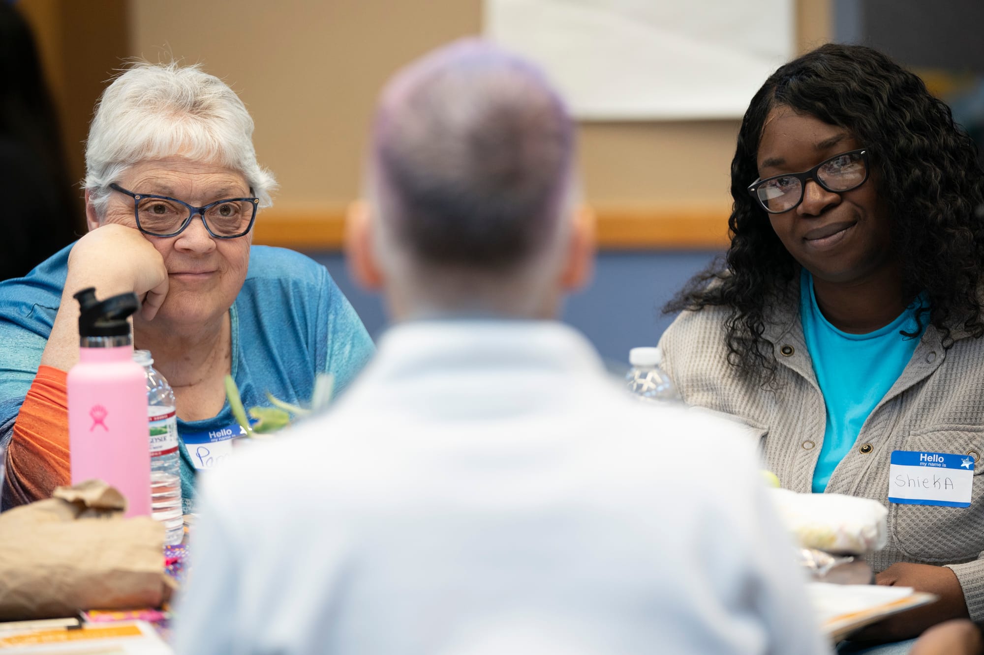 Two women face another woman, whose back is visible, during a moderated dialogue. The woman on the left has her hand on her cheek with a look of intent listening on her face. She wears blue glasses and a blue and orange shirt. She has gray hair. The woman on the right is also listening closely. She wears a light blue shirt under a gray button-down shirt. She has shoulder-length black, curly hair, and she wears black glasses. 