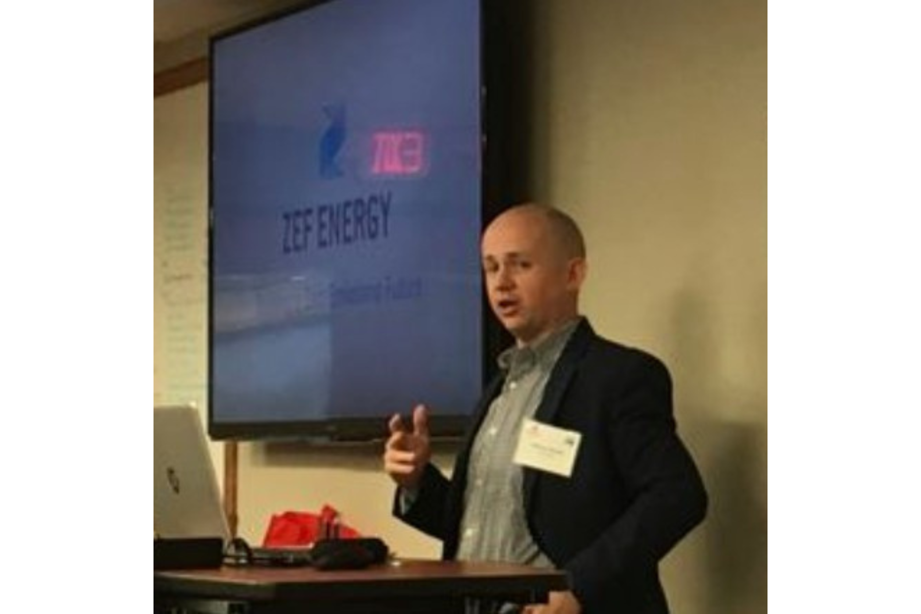 A man gives a presentation. He wears a dark suit jacket over a green button-down shirt. A projection screen is visible behind him with the words "ZEF Energy" on it.