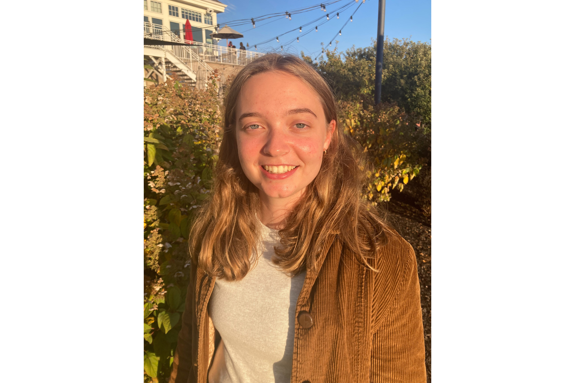 A young woman smiles for a photos. She wears a gray shirt underneath a light brown corduroy jacket. Her light brown hair is down. She's standing outside in front of some bushes.
