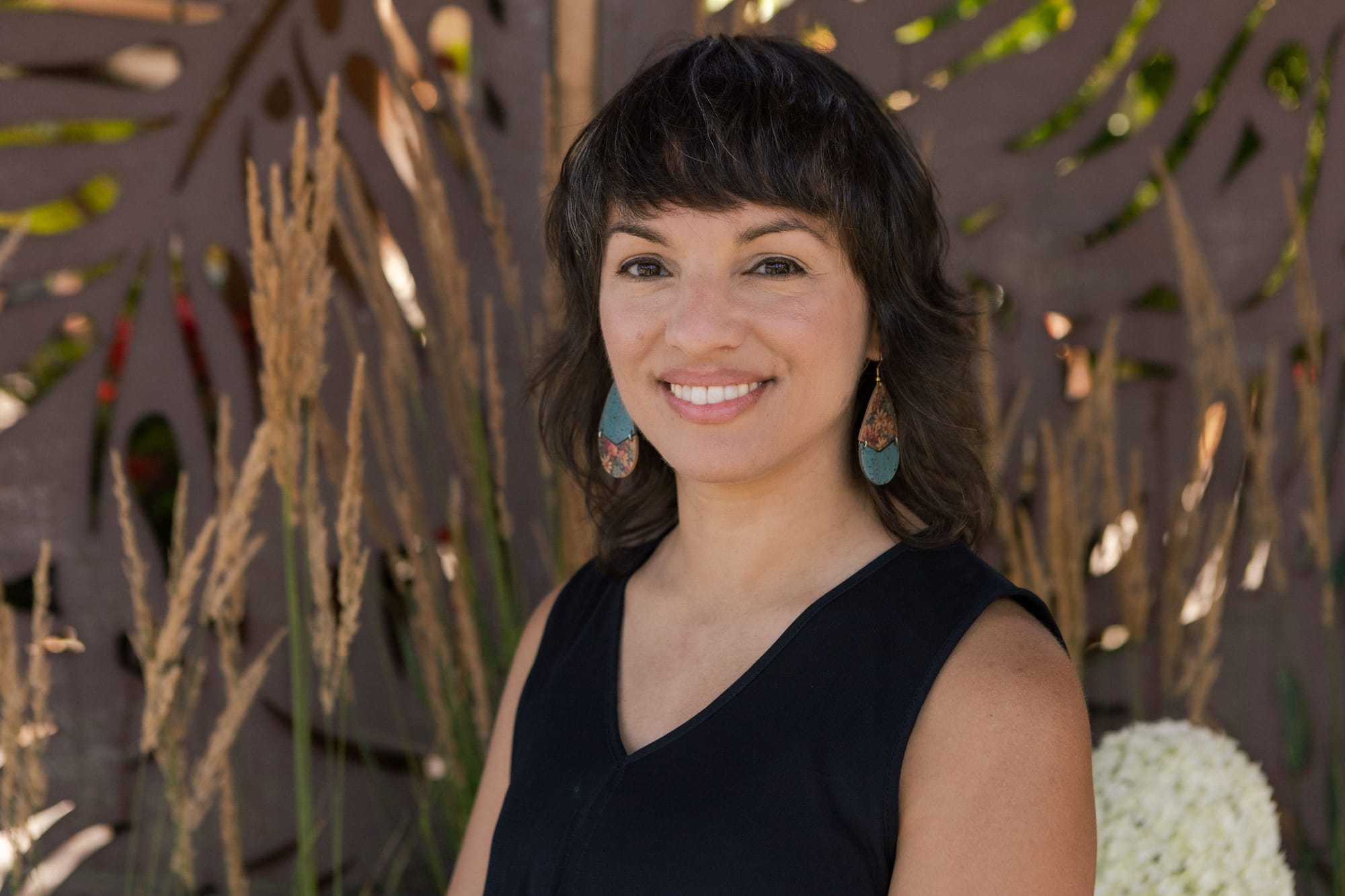 A woman smiles for a photo outside. She wears a black tank top. She has brown, wavy hair with bangs. She wears dangly earrings.