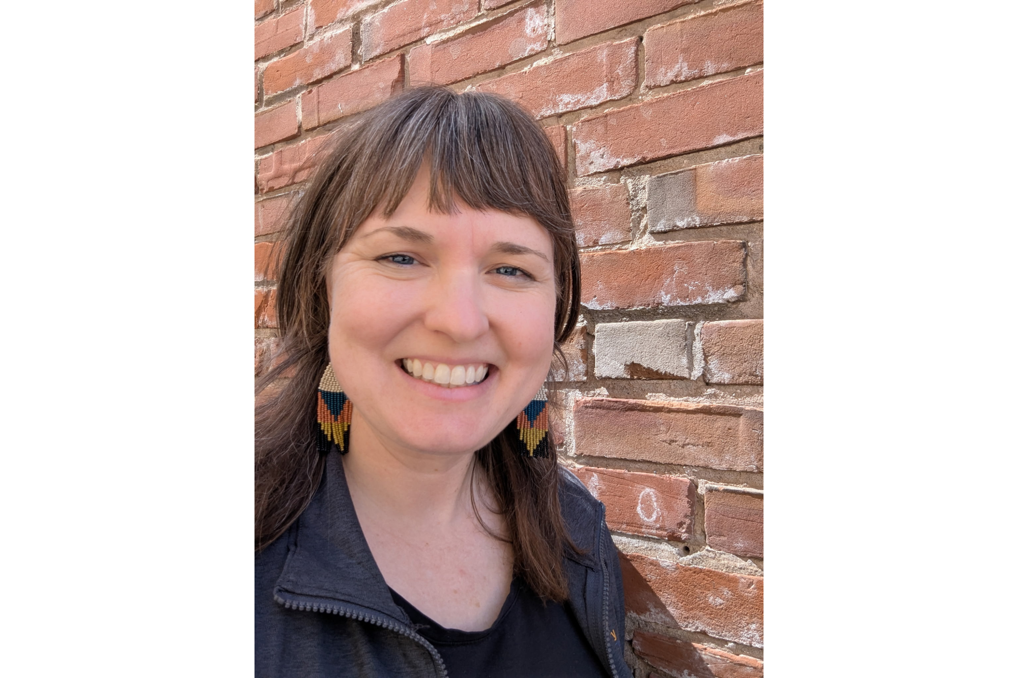 A woman smiles in front of a brick wall. She wears a black zip-up jacket over a black t-shirt. She has brown hair that falls around her shoulders and bangs. She wears beaded, multicolored earrings.