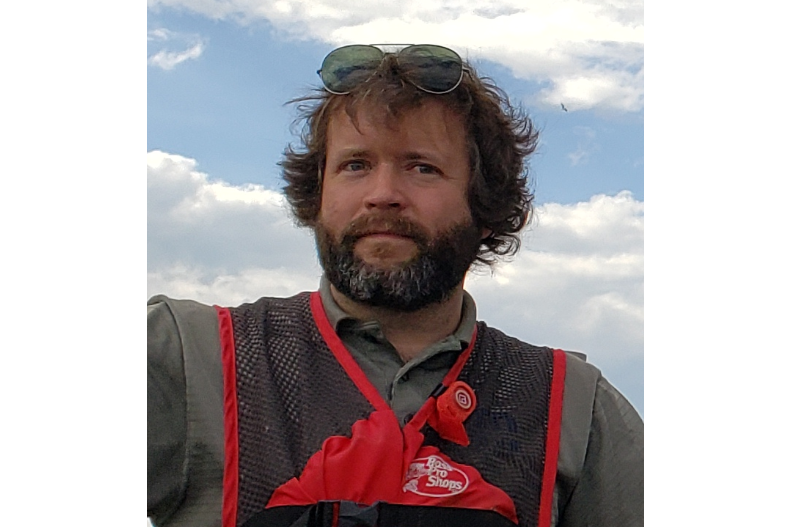 A white man is seen in a photo. He has hair long enough to be ruffled by the wind. His hair and beard are brown. His beard has flecks of gray in it. He wears sunglasses on the top of his head, so his eyes are visible to the camera. He wears a greenish gray shirt under a red and brown Bass Pro Shops fishing vest.