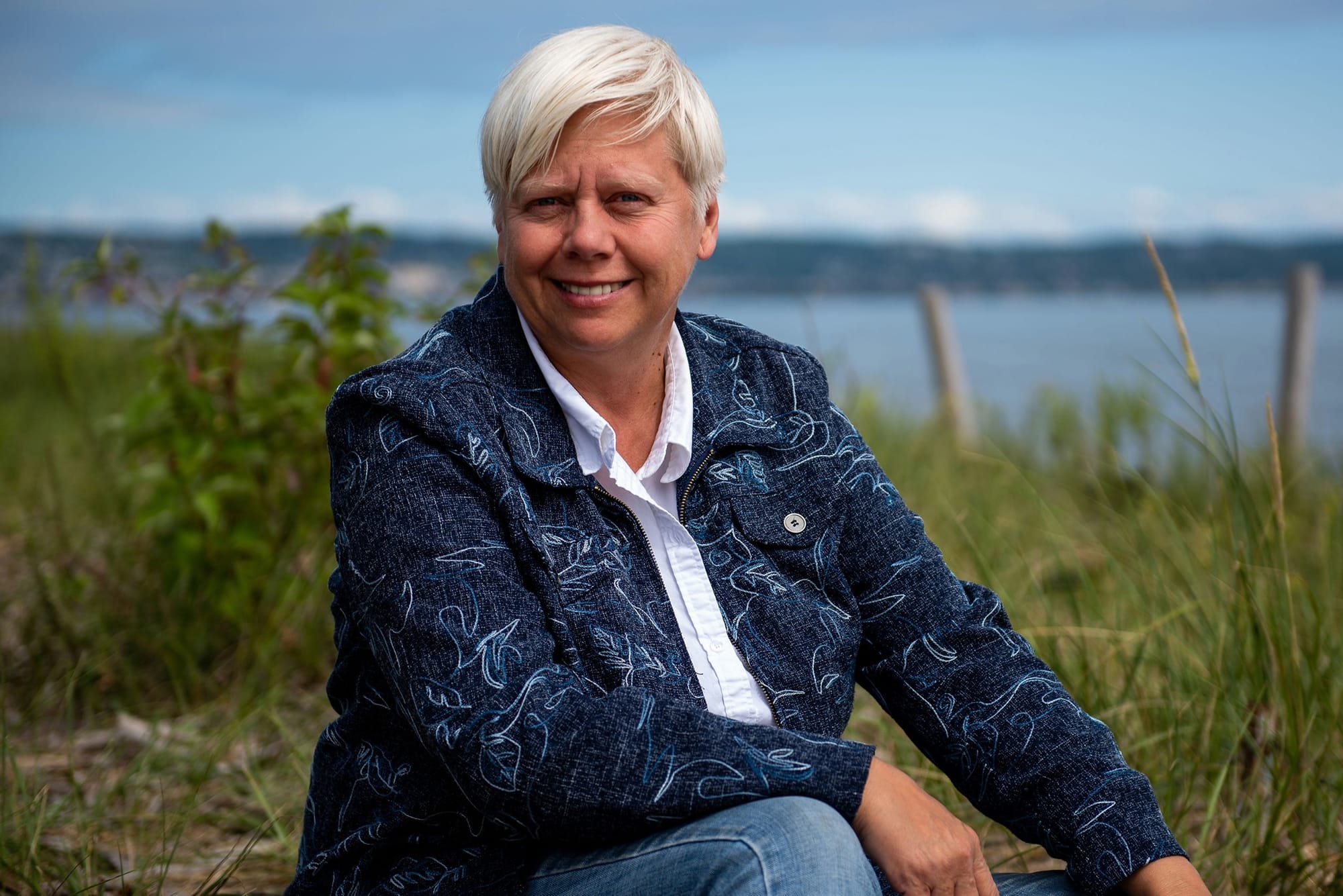 A white woman sits on the beach with tall grass behind her. She has short, white hair. She wears a white button-down shirt under a dark blue jacket and jeans.