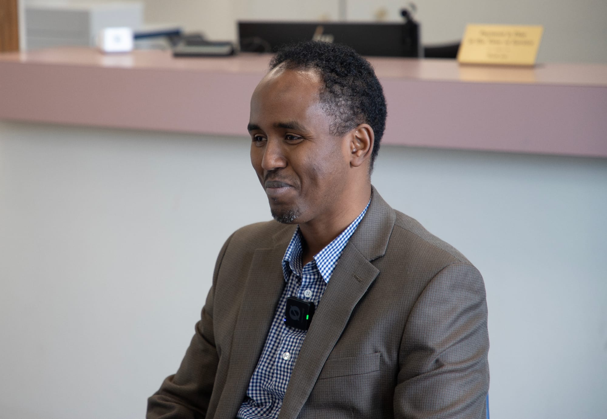 A man smiles during an interview. He looks to the left of the frame. He wears a blue striped button-down shirt under a gray suit jacket. He has short, black hair.