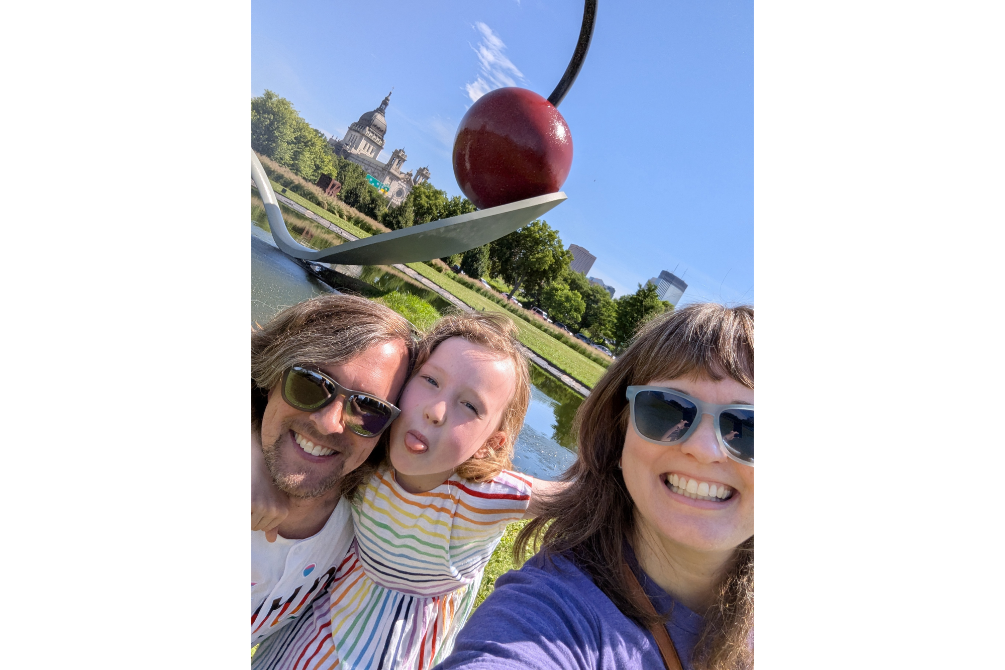 A family of three poses for a portrait at the Minneapolis Sculpture Garden in front of the spoon with the big cherry. The man has shoulder-length brown hair with gray streaks in it. He wears sunglasses and smiles. In the middle is their daughter, who sticks out her tongue for the photo. She wears a white dress with stripes the color of the rainbow. A woman is on the right with shoulder length brown hair and bangs. She smiles and wears sunglasses. The day is sunny with blue skies visible behind the cherry. 