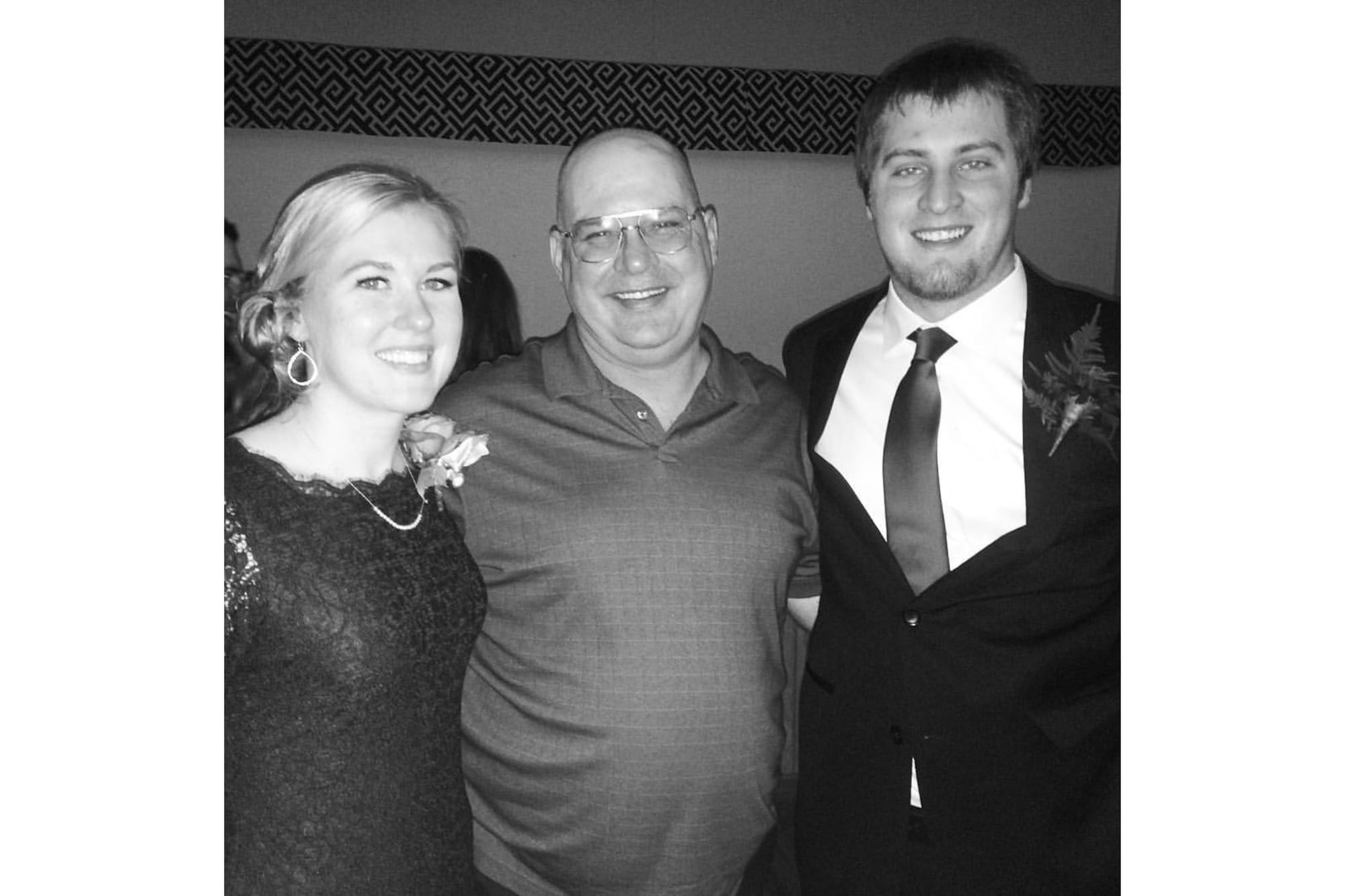 A young woman stands next to a bald man with glasses wearing a dress shirt and a young man wearing a suit. The image is black and white. 