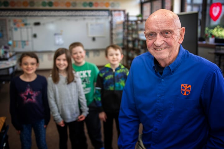 Man stands in a classroom in the foreground with children smiling in the background.