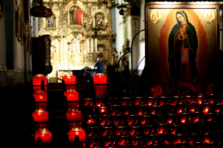 Red prayer candles are lit with an image of Mary in the background at a Catholic church. 