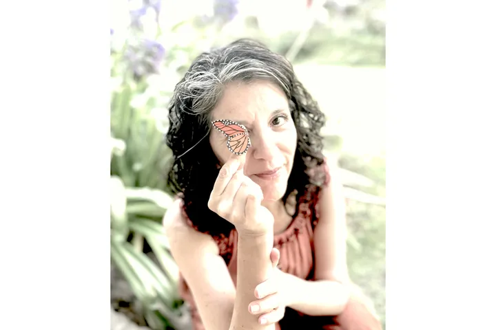 Una mujer sostiene un ala de mariposa monarca sobre uno de sus ojos mientras mira a la cámara. Lleva un vestido naranja quemado. Tiene el pelo oscuro y rizado. 