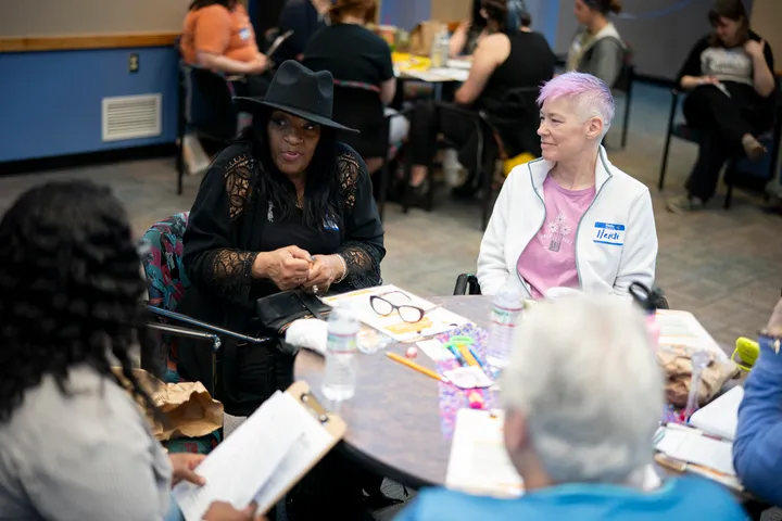 Five women sit around a table. We see three of their backs, and the faces of two. One woman, wearing a black shirt and a black hat, speaks. A woman next to her looks at her while she talks.