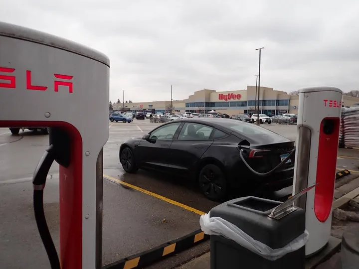 A black Tesla Model 3 is connected to a charger at a Hy-Vee in Rochester, Minn. The Hy-Vee store is visible in the background. It's an overcast day. 