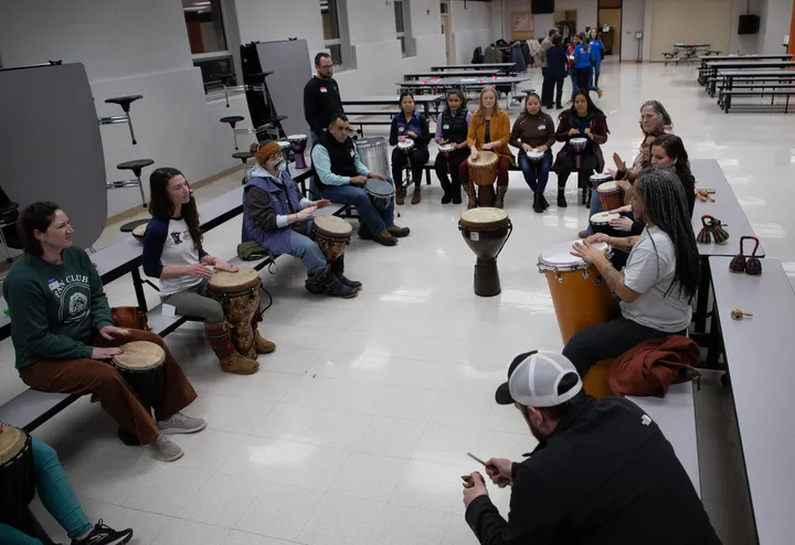 A group of people participate in a drum circle at a community center. They sit on cafeteria table benches with drums between their knees. 