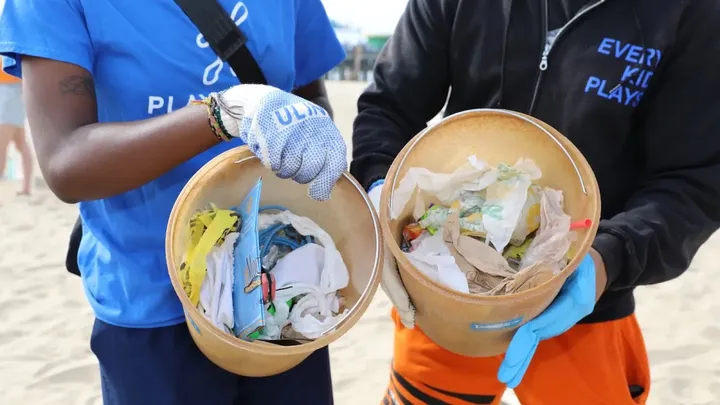 Two people hold buckets of garbage they collected on a beach. The image does not show their faces. You see their hands holding the buckets. They're wearing gloves on their hands. 